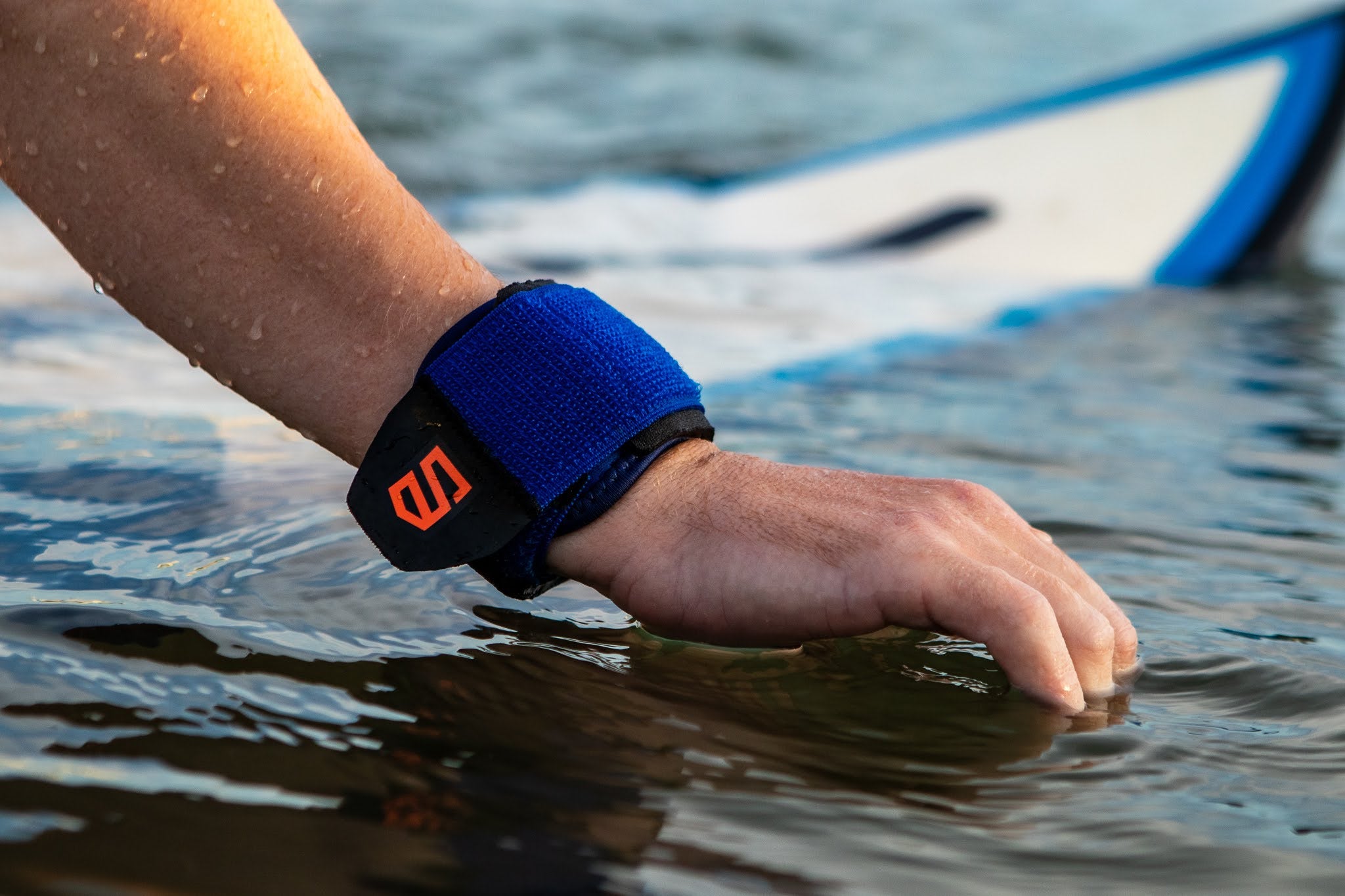 Person's hand wearing a blue wristband with an orange logo, touching water with a surfboard in the background.